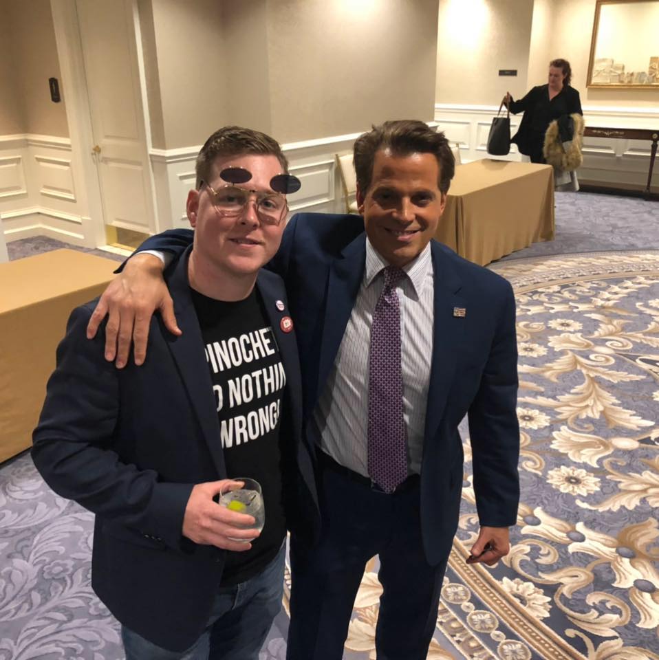 A photograph of two men posing together in a carpeted ballroom or lobby. The man on the left wears a blue blazer over a black t-shirt that reads 'PINOCHET DID NOTHING WRONG' and holds a drink. The man on the right, identified as Anthony Scaramucci, wears a blue suit and tie and has his arm around the other man.