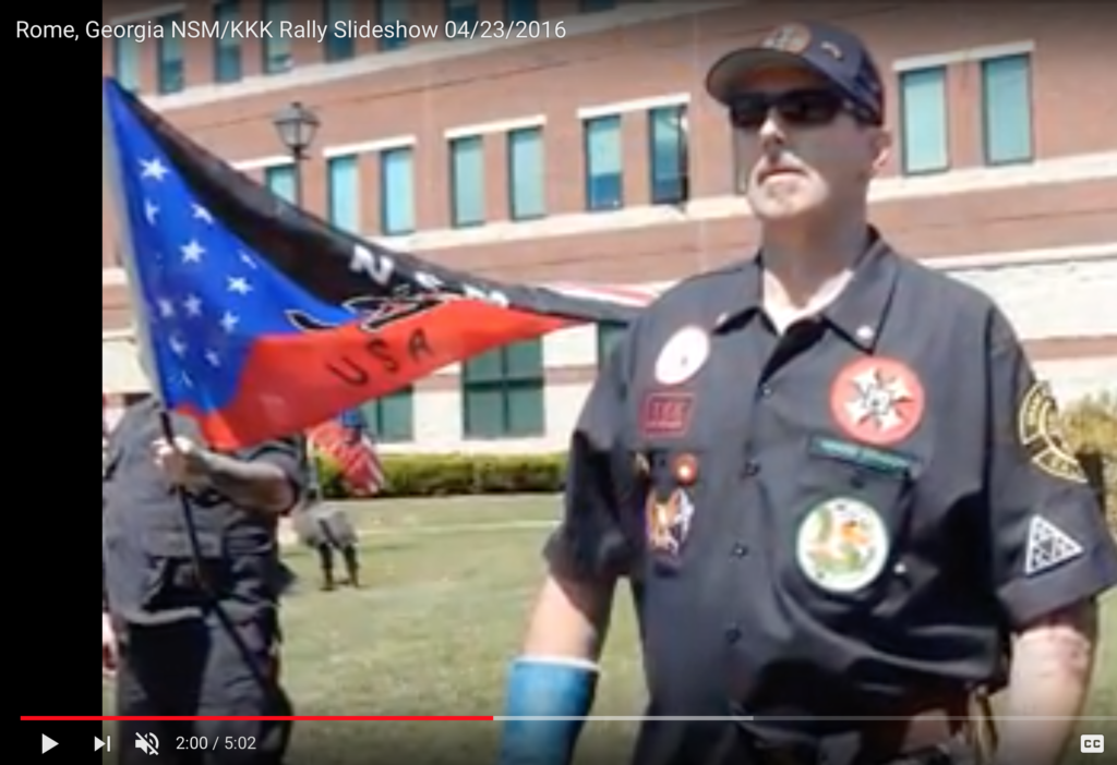 A screenshot from a video titled 'Rome, Georgia NSM/KKK Rally Slideshow 04/23/2016' showing a man in a black uniform with white supremacist patches, including a KKK cross and NSM logos, standing in front of a brick building.