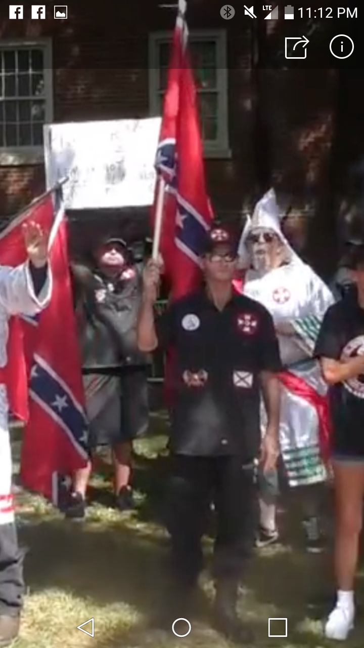 A mobile screenshot showing a group of white supremacists at a rally. Two individuals are dressed in traditional white KKK robes and hoods, while others wear black shirts with extremist patches. They are holding Confederate flags and a sign in front of a brick building.