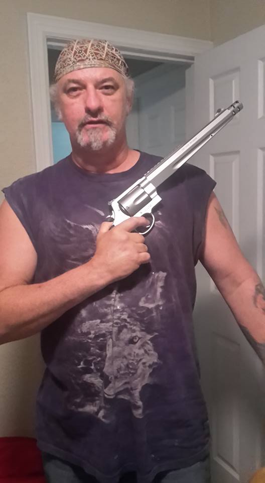 A Caucasian man with a goatee and a patterned skull cap holding a large silver revolver, wearing a sleeveless wolf t-shirt.