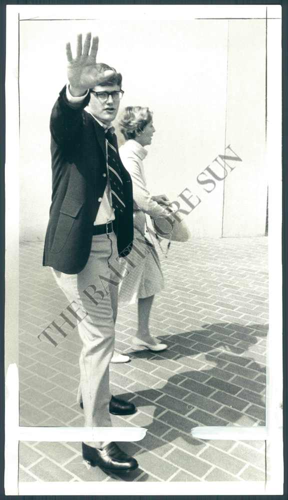 A black and white archival photograph from The Baltimore Sun showing a young man with dark hair and glasses wearing a suit jacket and tie, holding his hand up towards the camera lens. A woman walks behind him on a paved surface.