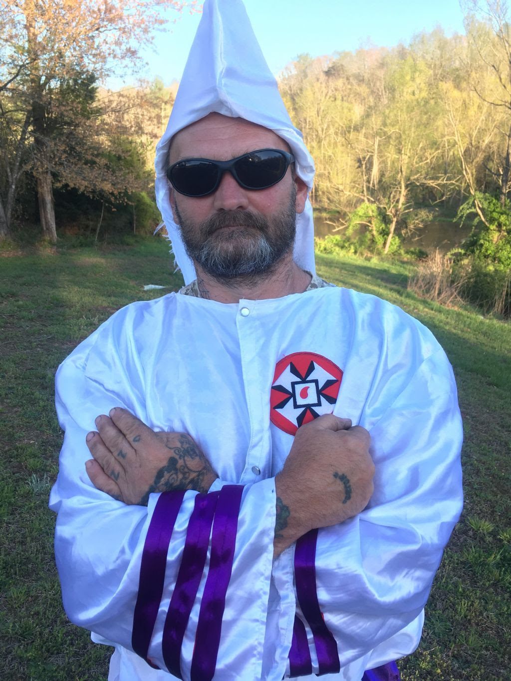 A photograph of a man in a white KKK robe with purple stripes on the sleeves and a red patch on the chest. He is wearing sunglasses and has a full beard and hand tattoos. He is standing outdoors with his arms crossed.
