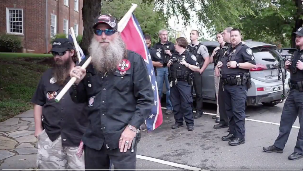 A photograph showing two men in black shirts with KKK-related patches, one holding a Confederate flag, standing in front of several police officers in a parking lot.