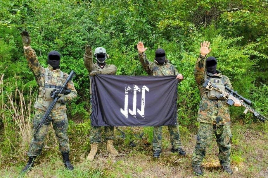 Four individuals in camouflage tactical gear and black balaclavas stand in a wooded area. They are performing a Nazi salute while holding a black flag with white extremist symbols. Two individuals are armed with rifles.