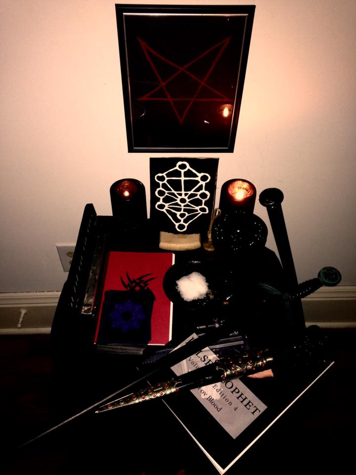 A photograph of a Satanic altar featuring an inverted pentagram on the wall, candles, occult symbols, a dagger, and a publication titled 'False Prophet'.