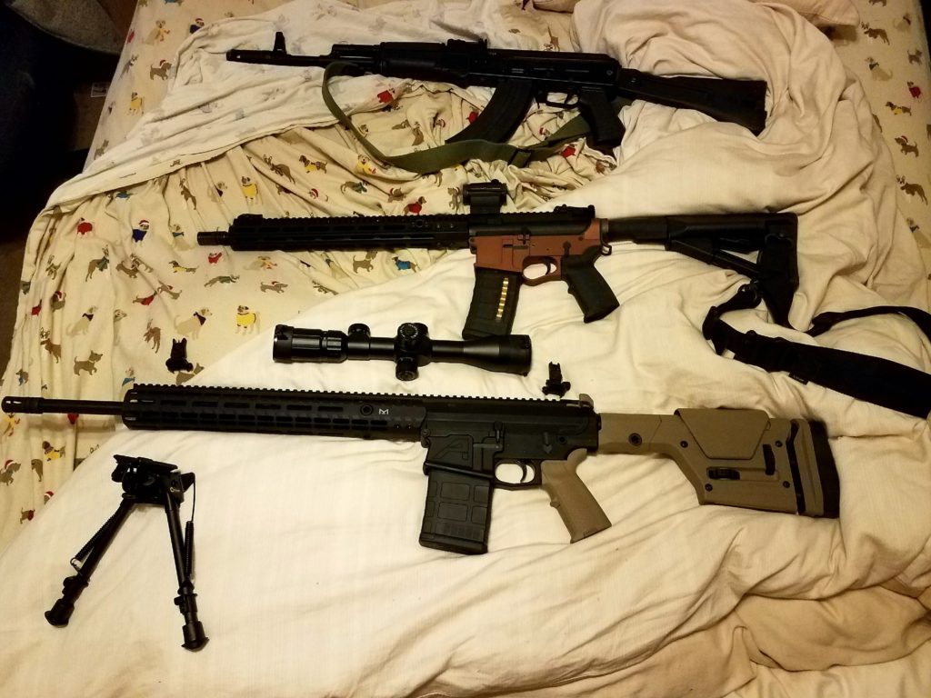 A photograph showing three rifles laid out on a bed with dog-patterned sheets. The top rifle is an AK-style firearm, the middle is an AR-15 style rifle with a reddish-brown receiver, and the bottom is a larger rifle with tan furniture. A scope and a bipod are also visible on the bed.