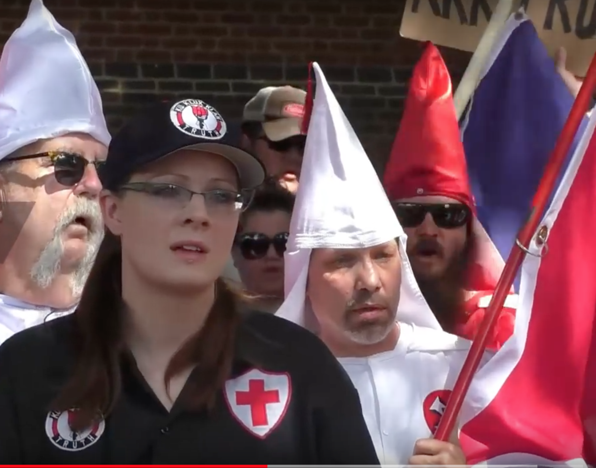 A group of individuals at a KKK-related gathering, featuring people in traditional white and red pointed hoods and a woman in a black shirt with KKK patches and a baseball cap.