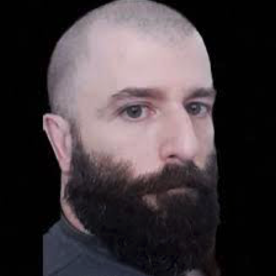 A close-up photograph of a Caucasian man with a shaved head and a thick, dark beard against a black background.