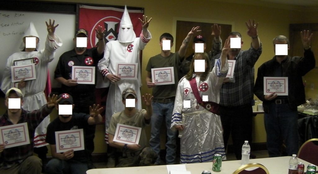 A group of individuals, some wearing Ku Klux Klan robes and hoods, posing with certificates and making hand gestures. Faces are obscured by white boxes.