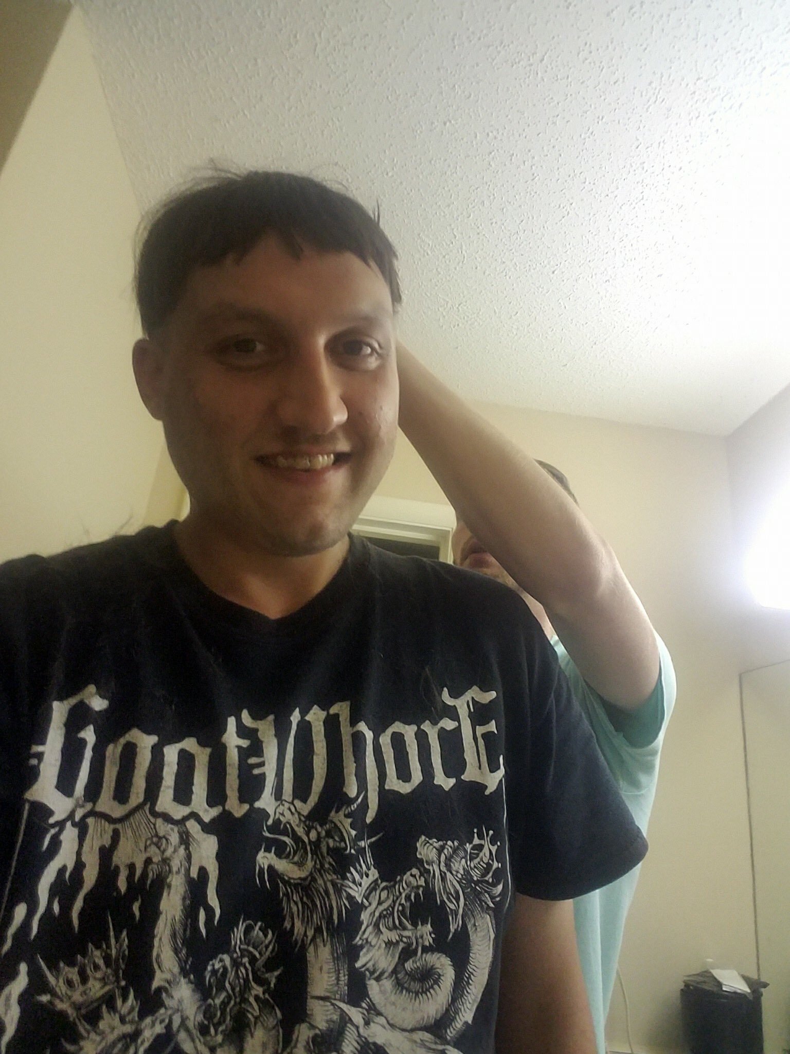 A photograph of a man with dark hair smiling, wearing a black 'Goatwhore' band t-shirt. Another person's arm is visible behind him.