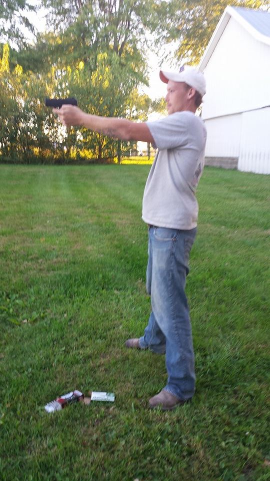 A man in a white t-shirt, blue jeans, and a white baseball cap aims a handgun in a grassy field next to a white building.