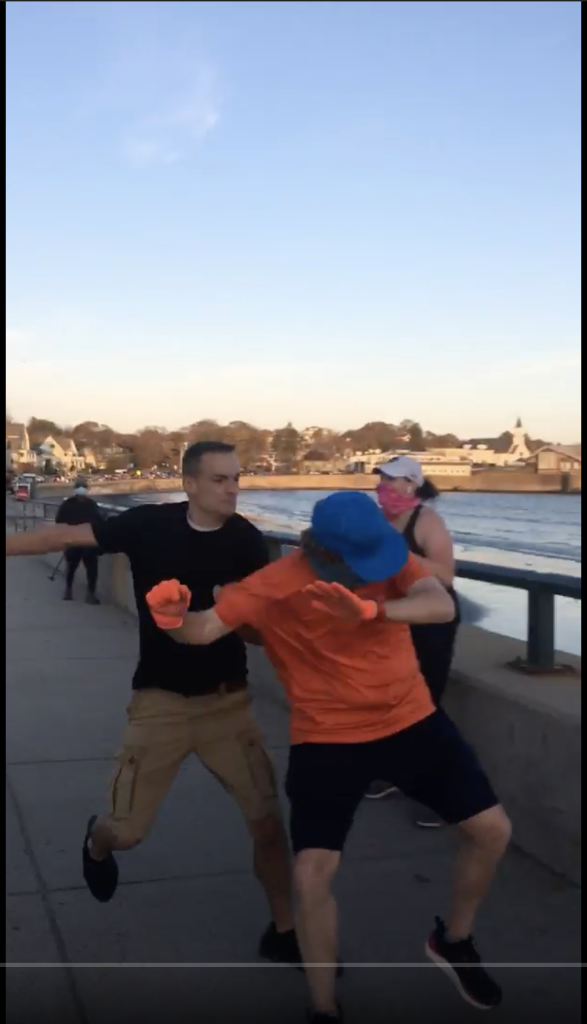 A screenshot of a physical altercation on a coastal boardwalk between a man in a black t-shirt and khaki shorts and another man in an orange t-shirt and blue hat.