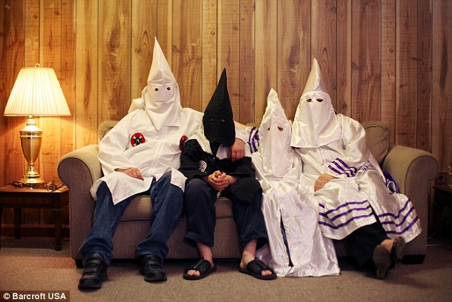 Four individuals wearing Ku Klux Klan robes and pointed hoods sit together on a couch in a wood-paneled room.