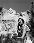 A black and white photograph showing a Native American man in traditional dress in the foreground with the Mount Rushmore National Memorial in the background.