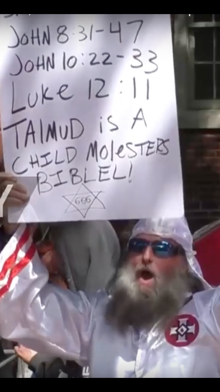 A man in a white KKK robe with a long grey beard and sunglasses shouts while holding an antisemitic sign with Bible verses and a Star of David.