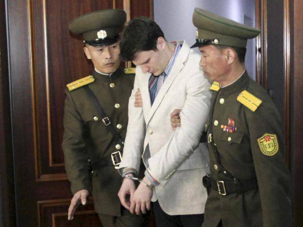 A photograph of Otto Warmbier, a young Caucasian man in a light suit and handcuffs, being escorted by two North Korean soldiers in olive green uniforms.