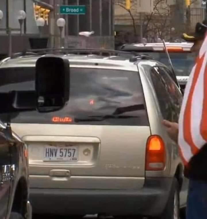 A silver minivan with Ohio license plate HNV 5757 stopped in traffic, with a person holding an American flag partially visible on the right side of the frame.