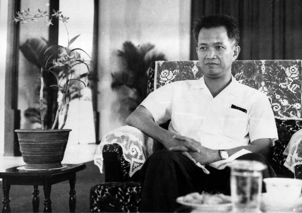 A black and white photograph of an Asian man with short dark hair sitting in an armchair, wearing a white short-sleeved shirt. A potted orchid is visible to the left.