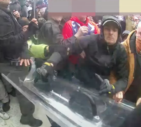 A photograph showing a chaotic scene at a protest or riot where individuals are pushing against a clear police riot shield. One man in a black hoodie and baseball cap is prominently visible in the center.