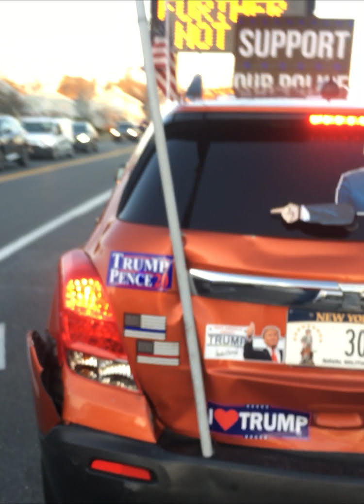 A photograph of the rear of an orange SUV covered in pro-Trump stickers and signs, including a 'Trump Pence 2020' sticker and an 'I ❤️ Trump' bumper sticker. The vehicle has a New York license plate and visible damage to the rear left bumper.