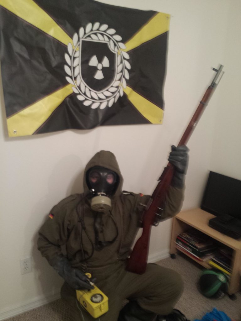 A person in a full olive-drab military or hazmat suit with a gas mask, holding a bolt-action rifle. They are positioned in front of an Atomwaffen Division flag, which features a radiation symbol inside a wreath on a black and yellow background.