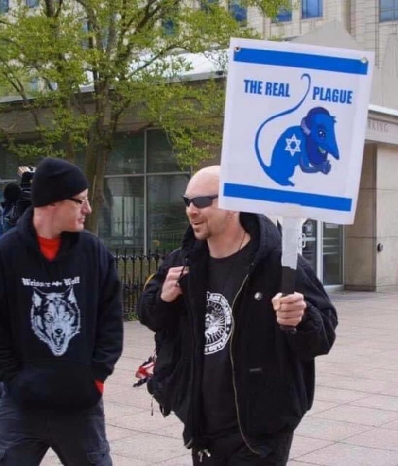 A photograph of two men at a protest. One man on the right is bald, wearing sunglasses and a black jacket, holding a sign with an antisemitic caricature and the words 'THE REAL PLAGUE'. The man on the left is wearing a black beanie and a hoodie with a wolf graphic.
