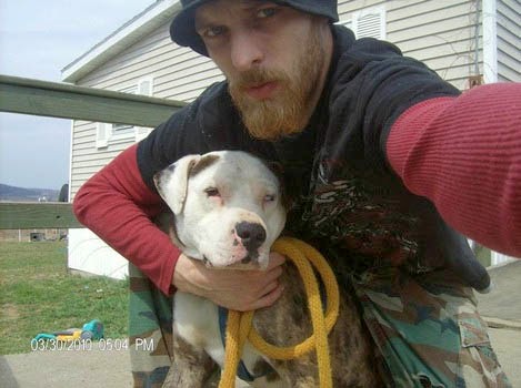 A selfie of a man with a full beard and a black beanie holding a white dog with brown spots. He is wearing a black t-shirt over red sleeves and camouflage pants, standing in front of a house.