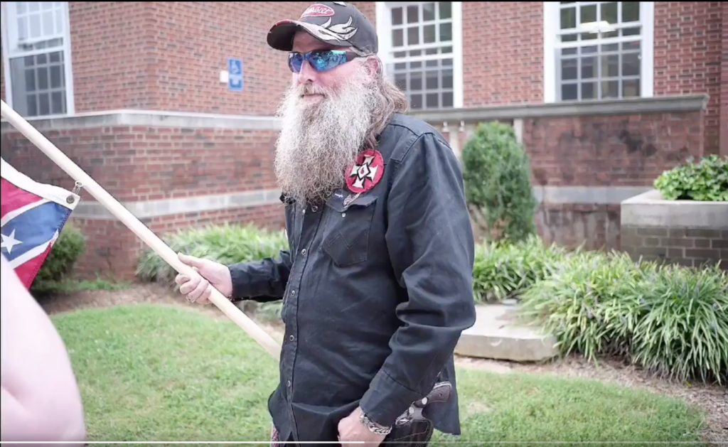 A photograph of a Caucasian man with a long, grey beard and a baseball cap, wearing a black shirt with a KKK patch and holding a Confederate flag in front of a brick building.