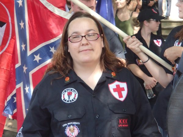 A woman with long brown hair and glasses wearing a black shirt adorned with Ku Klux Klan (KKK) patches, including 'Loyal White Knights' and 'KKK Life Member', standing in front of a Confederate flag.