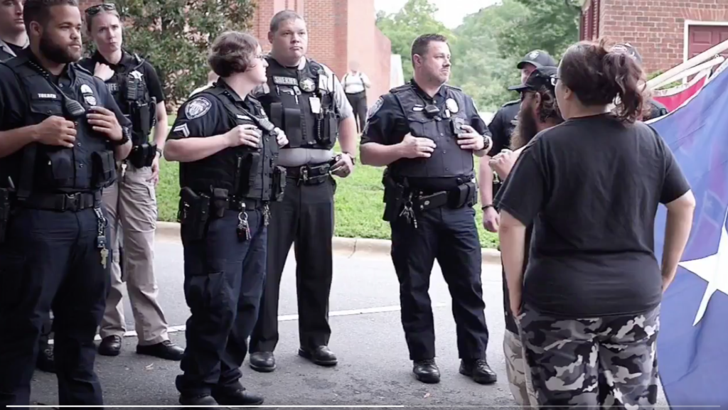 A group of police officers in uniform standing on a street, facing a civilian wearing camouflage pants and a black shirt next to a Confederate flag.
