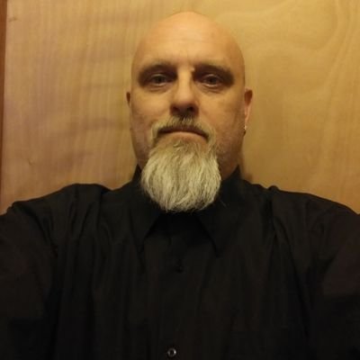 A close-up photograph of a bald Caucasian man with a long grey goatee, wearing a black button-down shirt against a wooden background.