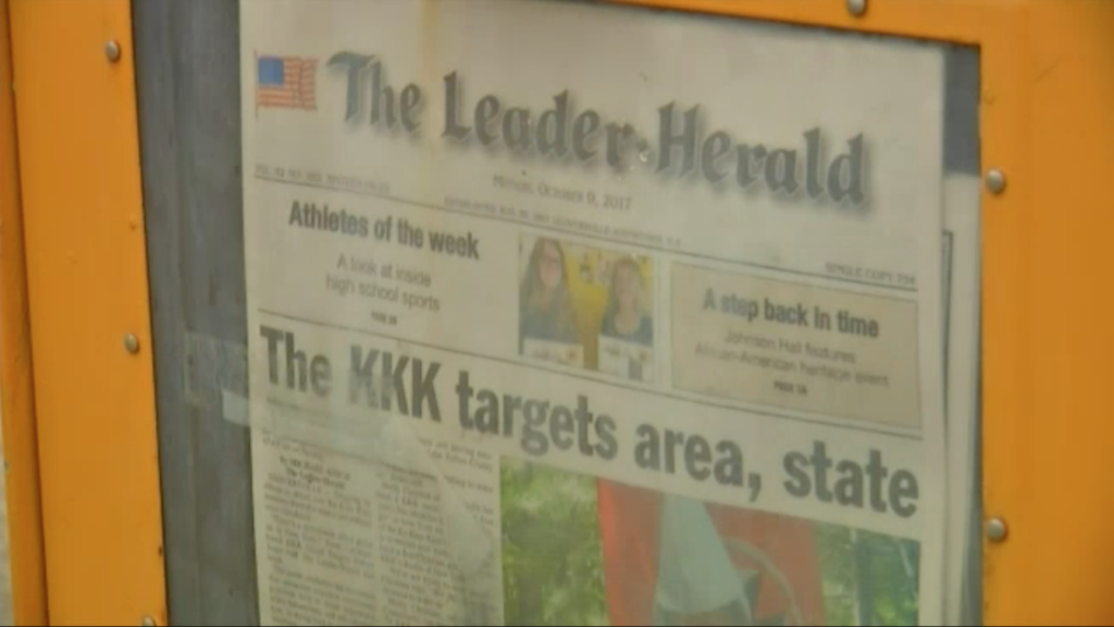 A photograph of a newspaper titled 'The Leader-Herald' inside a yellow display box. The front-page headline reads 'The KKK targets area, state'.