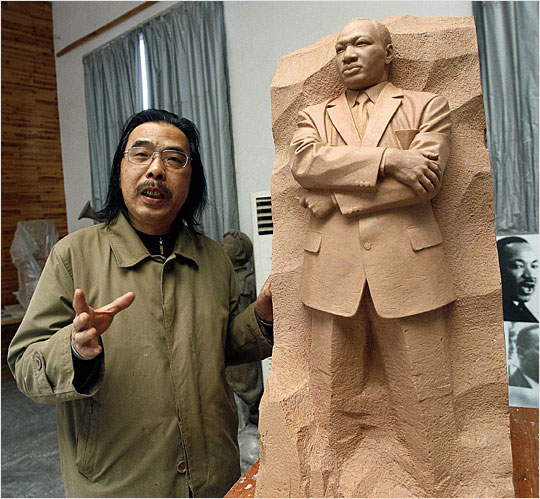 A photograph of a man with long black hair and glasses standing next to a clay model of a Martin Luther King Jr. monument.