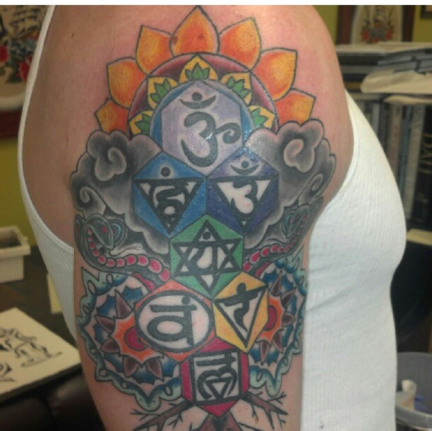 A close-up photograph of a large, colorful tattoo on a person's upper arm, featuring various spiritual and geometric symbols including Sanskrit characters and a Star of David.