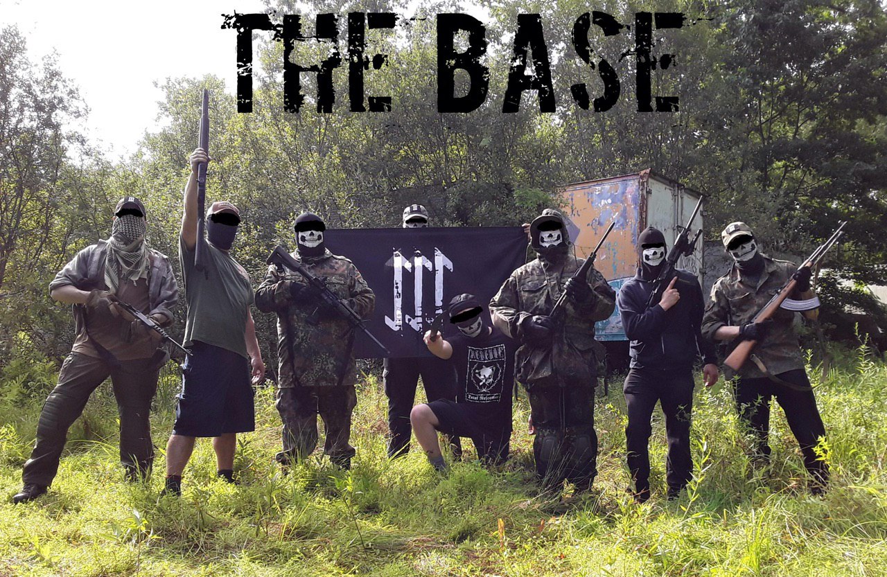 A group of eight individuals in tactical gear and masks, some holding firearms, posing in a wooded area in front of a black flag with a white symbol. The text 'THE BASE' is written across the top of the image.