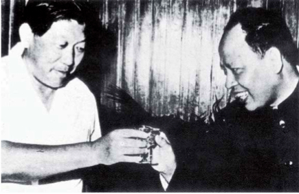A black and white historical photograph of two Asian men clinking glasses in a toast. The man on the right appears to be Pol Pot, and the man on the left is likely a Chinese official, possibly Hua Guofeng.