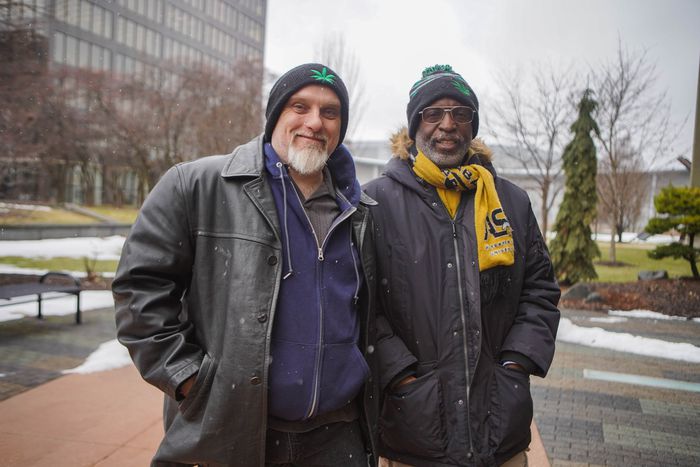 A photograph of two men standing together outdoors in a snowy urban environment. On the left is a Caucasian man with a white goatee wearing a black leather jacket and a beanie with a green leaf logo. On the right is an African American man wearing a black parka, a yellow scarf, and a matching beanie.