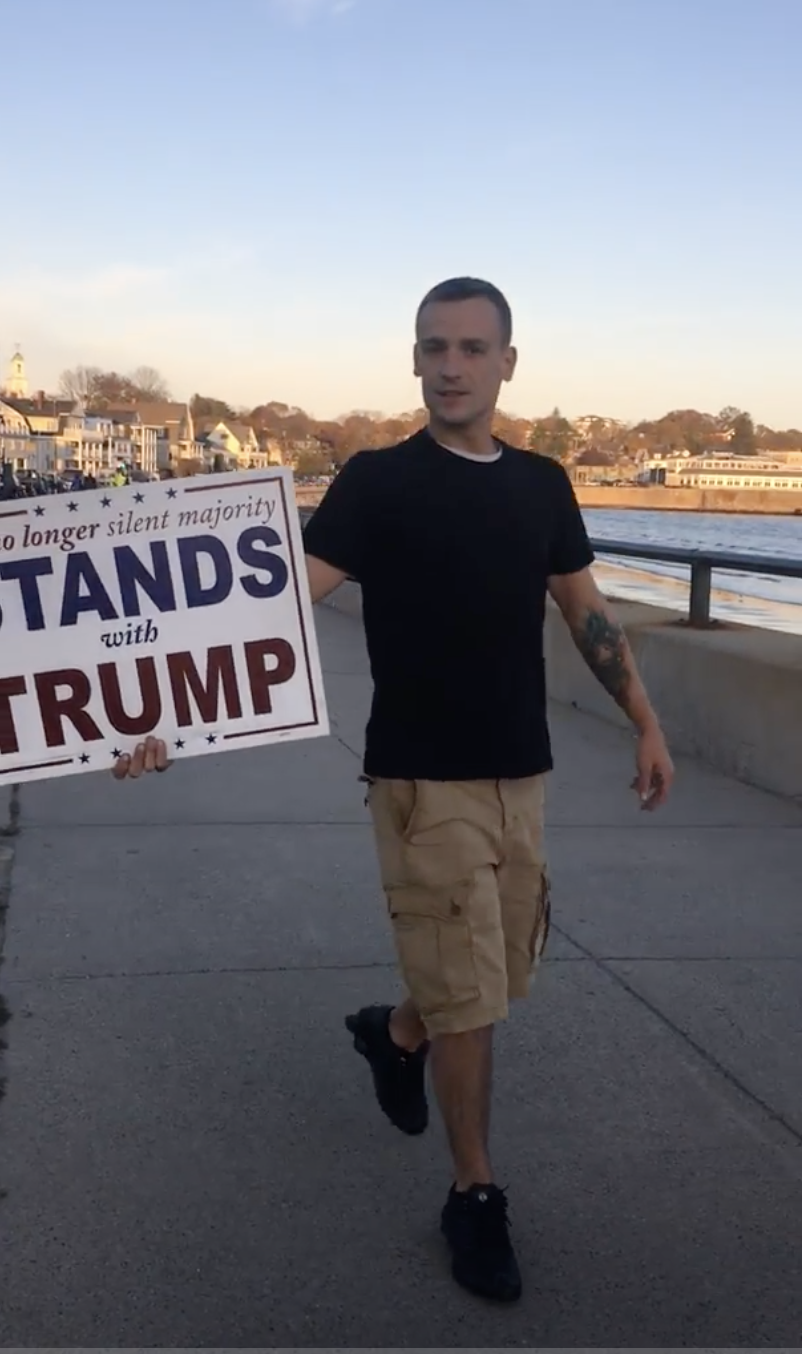 A man with short dark hair, wearing a black t-shirt and khaki cargo shorts, walks along a coastal boardwalk while holding a large sign that reads 'no longer silent majority STANDS with TRUMP'.