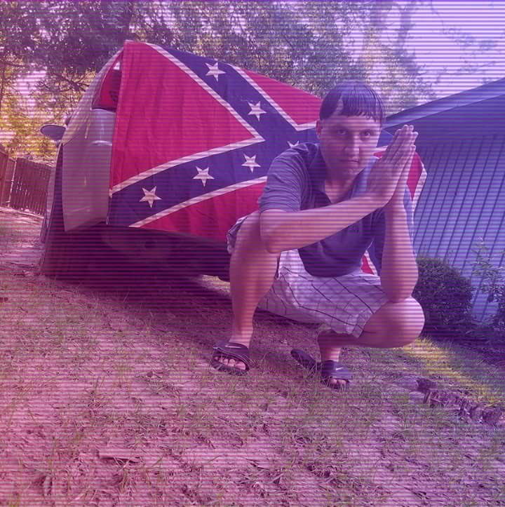 A photograph of a young man with dark hair crouching in front of a car with a Confederate flag draped over it. He has his hands pressed together in a prayer gesture. The image has a purple tint and horizontal scan lines.