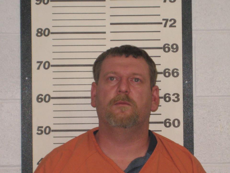 A mugshot of a Caucasian man with short brown hair and a goatee, wearing an orange jumpsuit, standing in front of a height chart.