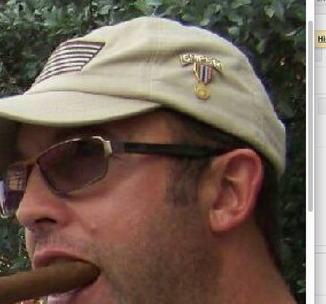 A close-up profile of a Caucasian man wearing a tan baseball cap with an American flag patch and a pin, dark sunglasses, and smoking a cigar.