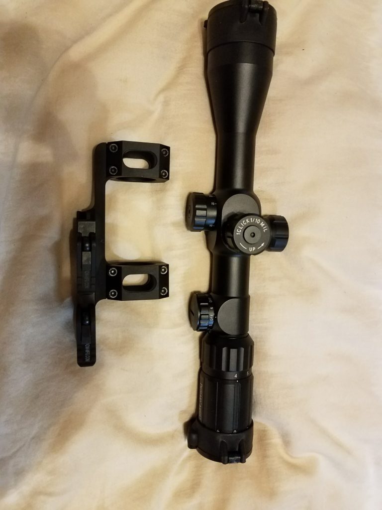 A black rifle scope and a scope mount lying on a light-colored fabric surface.