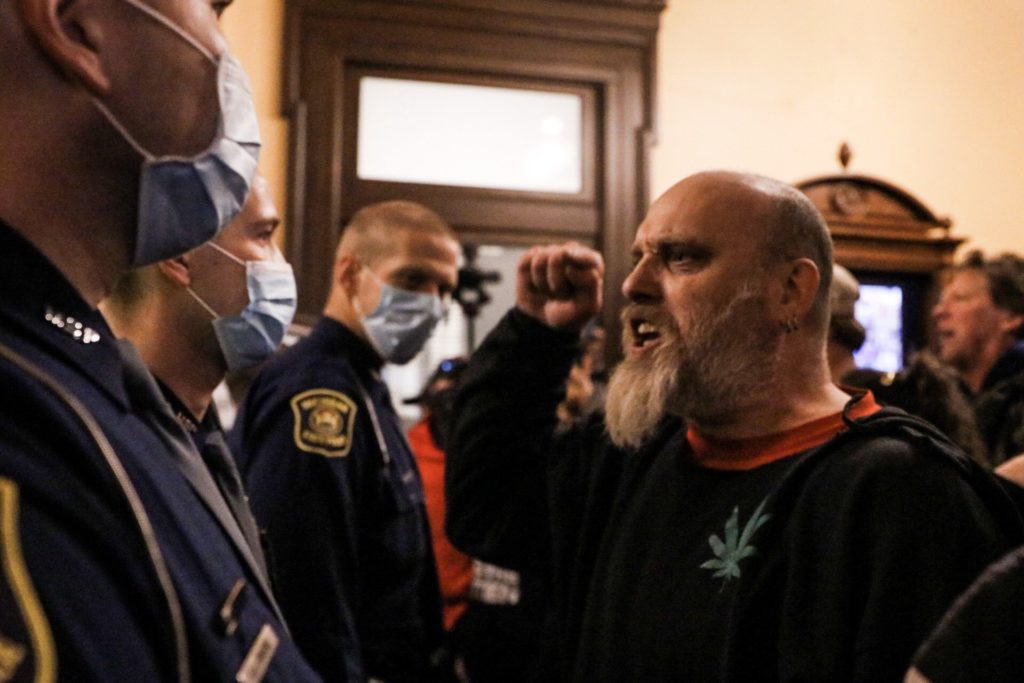 A bald Caucasian man with a long grey beard shouting and raising a clenched fist at masked police officers inside a building.