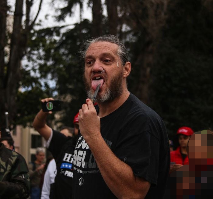 A photograph of a man with long hair and a full beard sticking his tongue out and touching it with a small blade. He is wearing a black t-shirt and is surrounded by people in an outdoor setting.