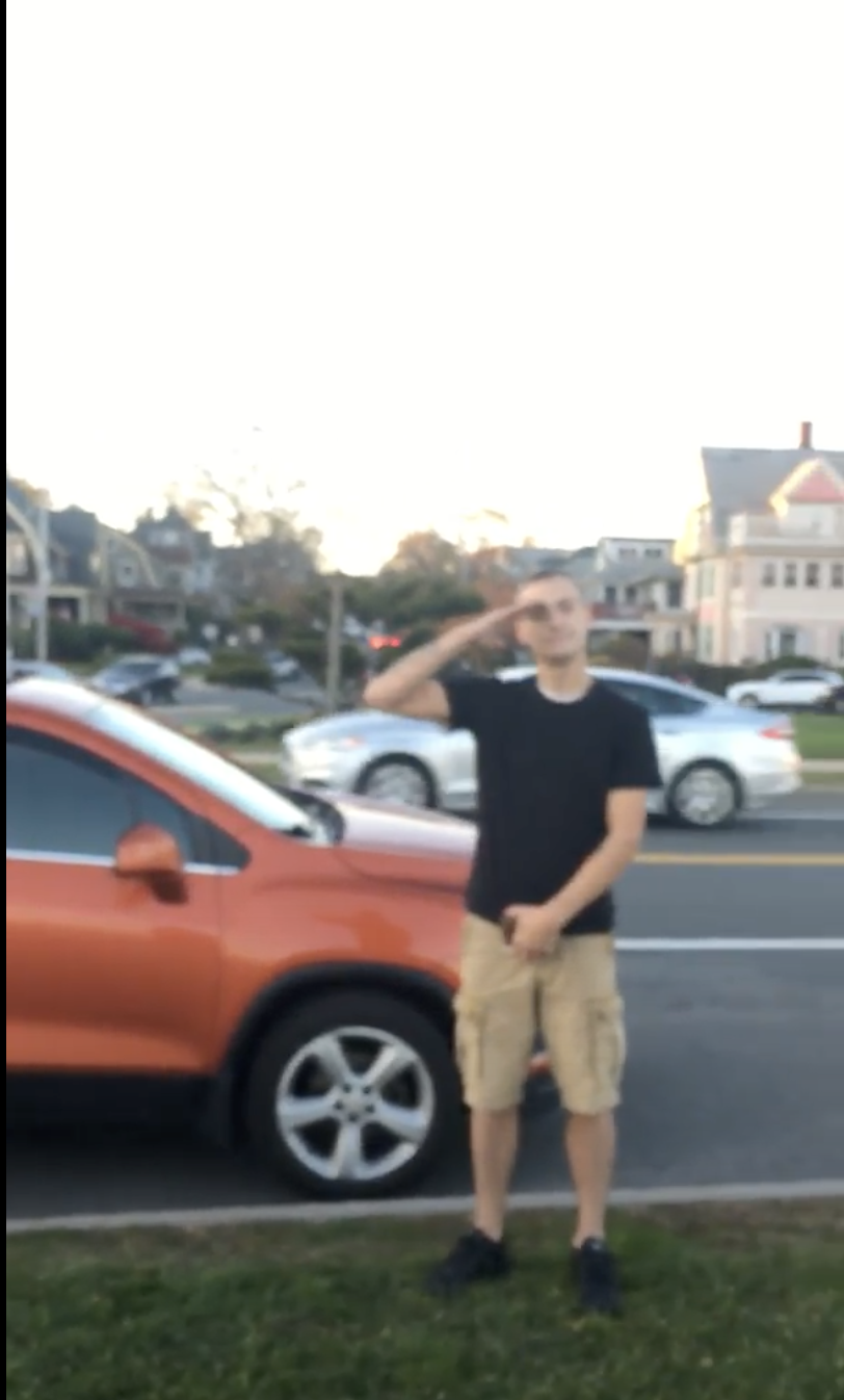 A blurry photograph of a man in a black t-shirt and khaki shorts saluting while standing next to an orange SUV on a residential street.