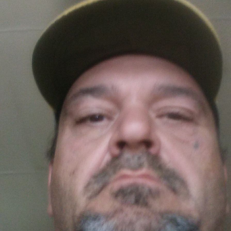 A low-angle close-up photograph of a Caucasian man with a goatee and mustache wearing a dark baseball cap.