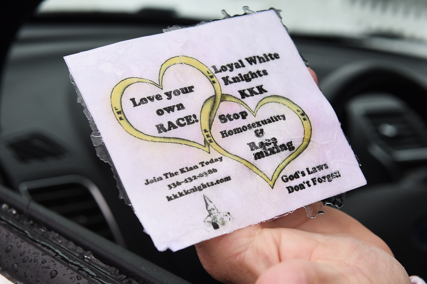 A hand holds a KKK recruitment flyer featuring two interlocking hearts and text that reads 'Love your own RACE!', 'Loyal White Knights KKK', and 'Stop Homosexuality & Race mixing'.