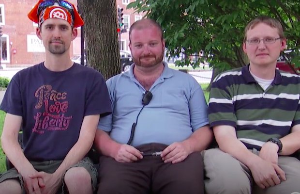 Three Caucasian men sitting on a park bench. The man in the center is heavy-set with a beard and short hair, wearing a light blue polo shirt. The man on the left wears a dark t-shirt and an orange bandana. The man on the right wears a striped polo shirt and glasses.