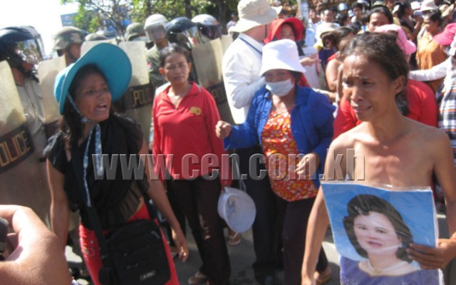 A photograph of a protest in Cambodia featuring several women in the foreground, one holding a portrait, facing off against a line of riot police with shields. A watermark for cen.com.kh is present.