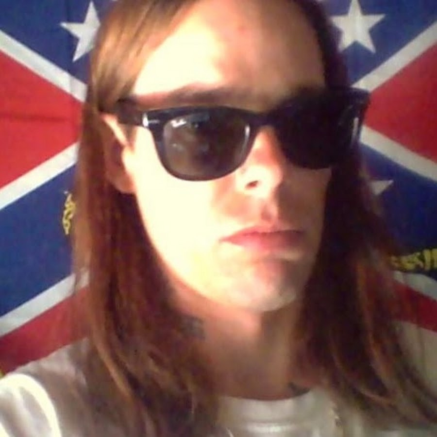 A Caucasian male with long brown hair and dark sunglasses posing in front of a Confederate flag.